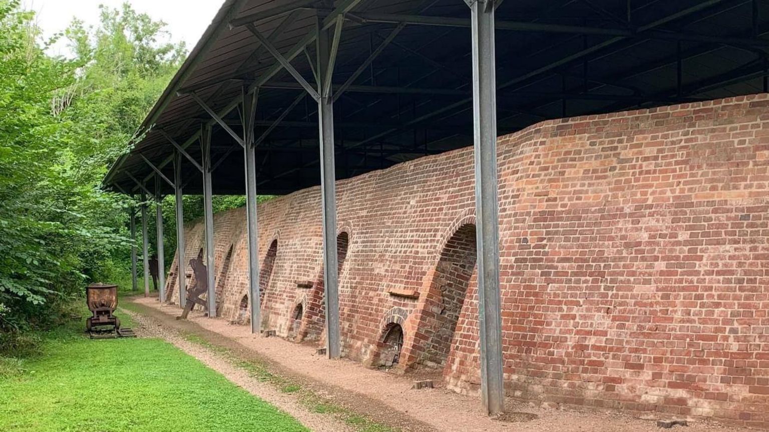 A large brick wall with multiple rounded doorways down its length. Above it, is a large roof which is supported by large metal beams in the ground. Leaning on one of the metal beams is a metal sculpture of a man.