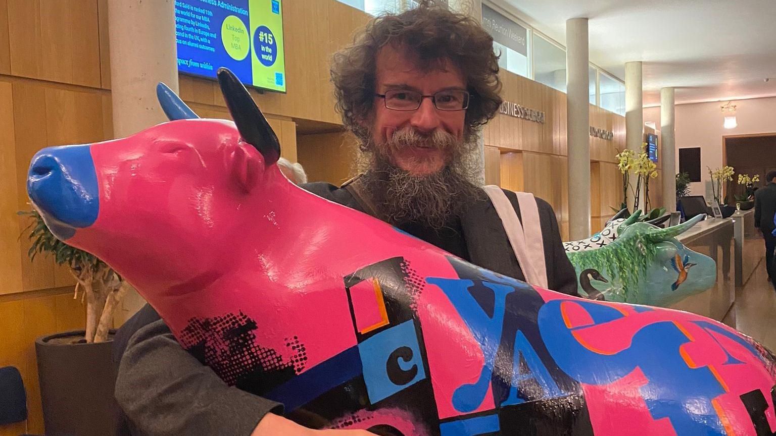 A man with curly hair and glasses smiling at the camera as he holds a pink blue and black coloured ox