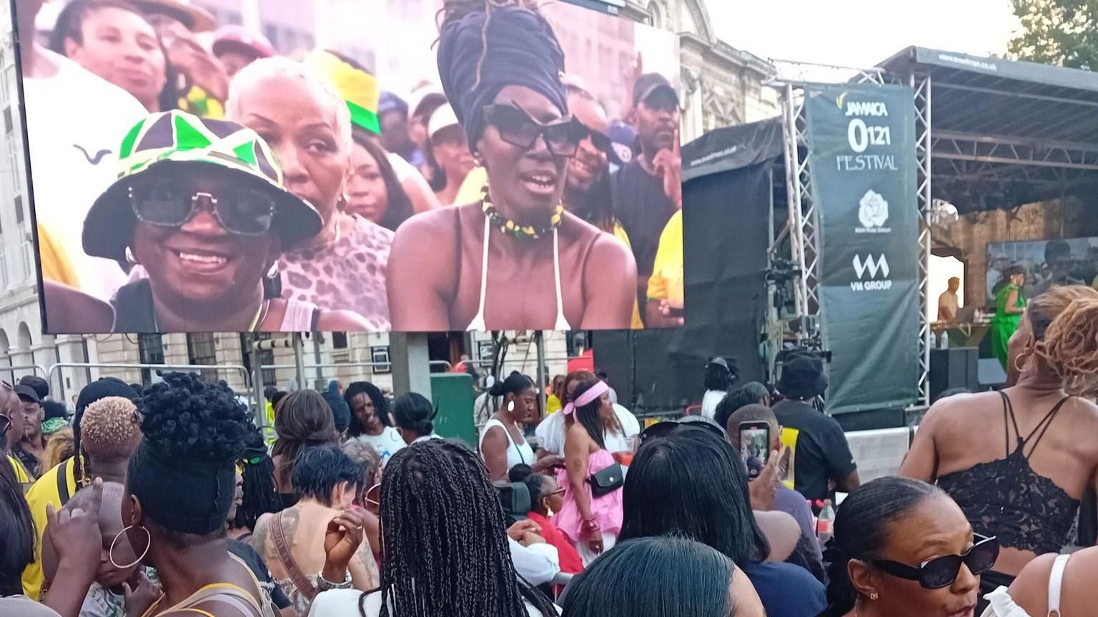 Black women are seen on a big screen in front of the heads of other people, some with long black hair, in front of a stage