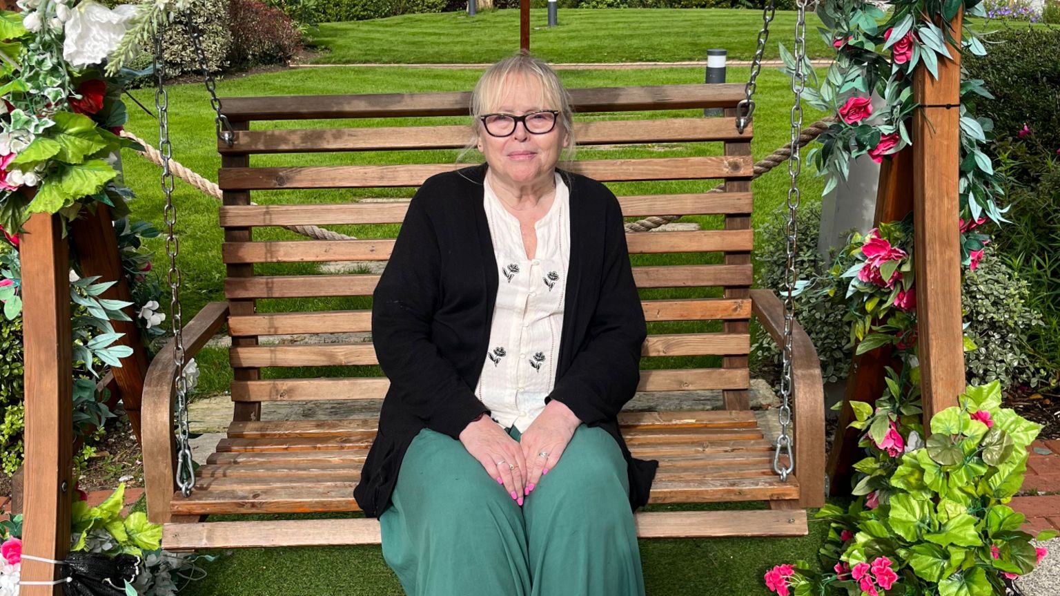Hazel Busby sitting on a wooden swing bench surrounded by flowers