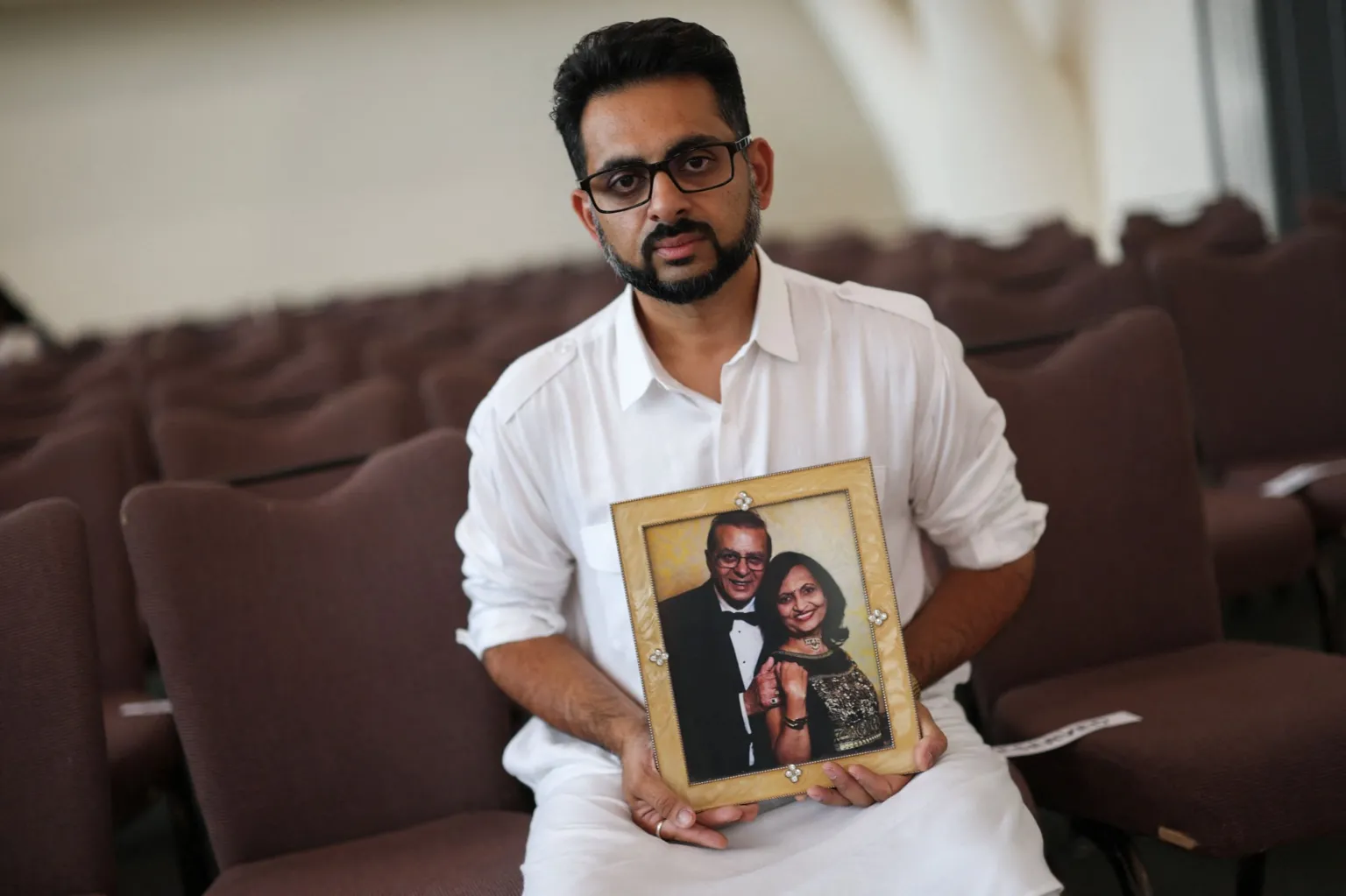 Reuters / Hannah Mackay A person in a white shirt sits among rows of empty auditorium seats, holding a framed photo of two people dressed in formal clothes. The background is softly out of focus, drawing attention to the person and the picture.