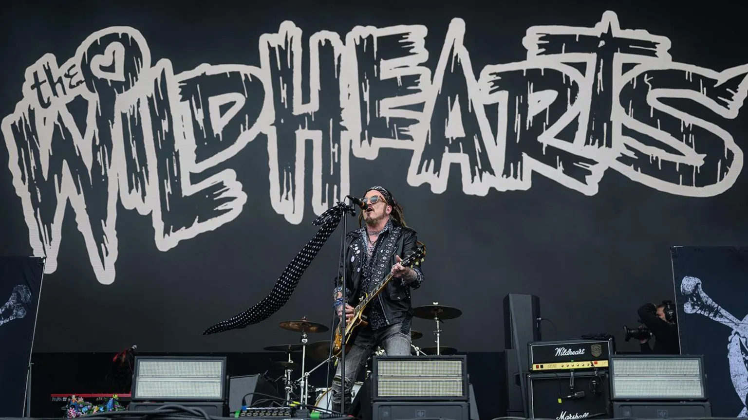 Getty Images A man in a black leather jacket, playing guitar on stage. He is singing into a microphone. Behind him is a black backdrop, with "The Wildhearts" written in white letters.