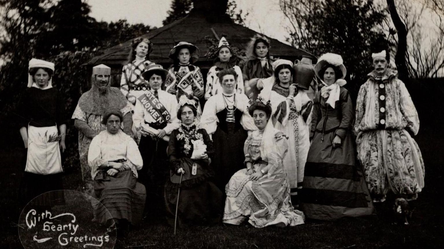 A black-and-white photo, possibly from the early 20th Century, of a group of 14 men and women wearing various fancy dress costumes, including a clown suit, knight's outfit and elaborate dresses.