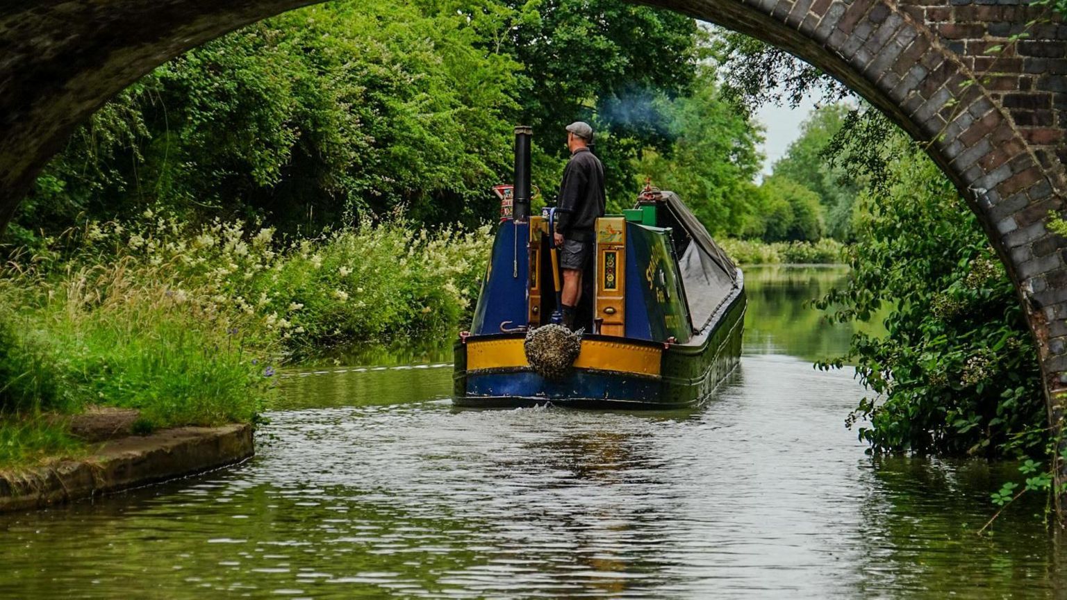 A blue and yellow narrowboat on the canal in Nuneaton, with a stone bridge in the foreground. A man in a cap and shorts is at the tiller 