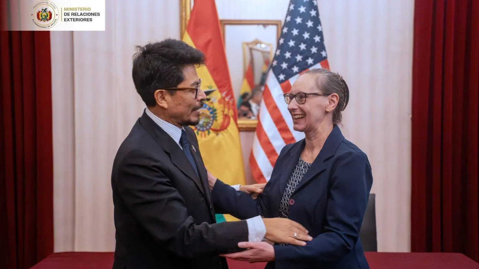Cancillería Bolivia A man in a suit and glasses, with short dark hair, holds the arms of a woman wearing a shirt and suit jacket and glasses, in front of the American and Bolivian flags.