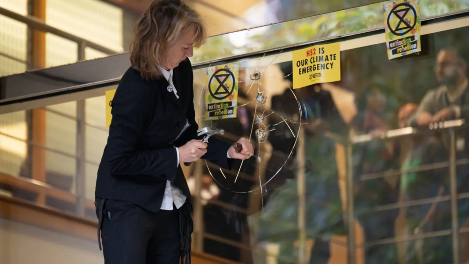 The image shows a woman in a dark jacket using a hammer-like tool to create spider web cracks in a large glass window or door. Yellow stickers with 