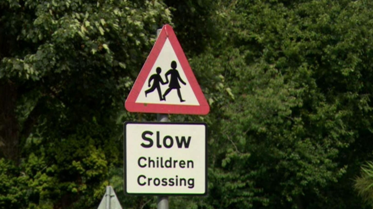 'Slow Children Crossing' red, white and black sign with green trees behind.