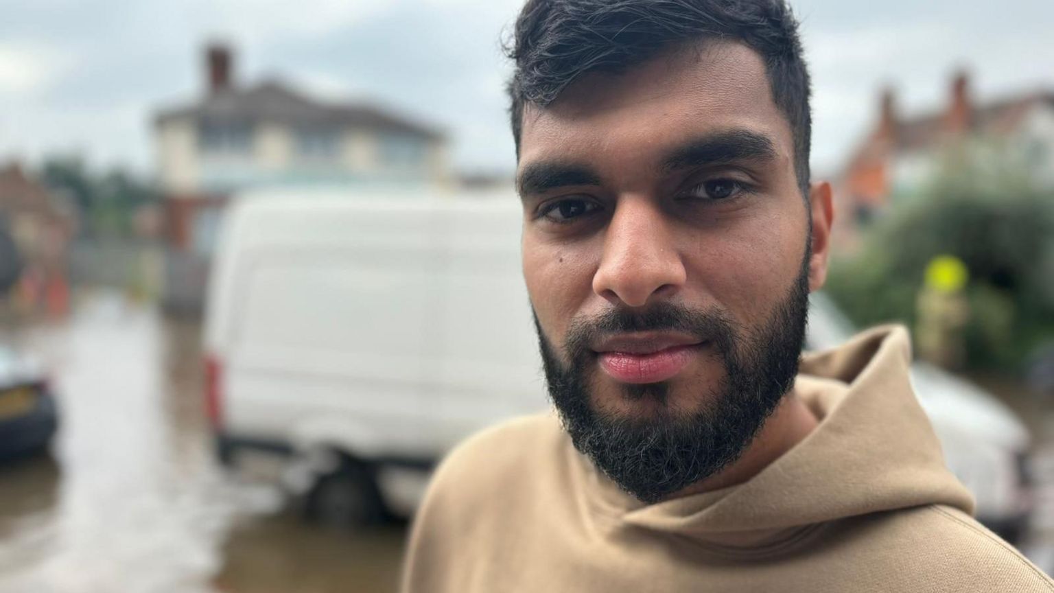 A man looks at the camera, with a blurred scene behind him showing a white van and a house in the distance.