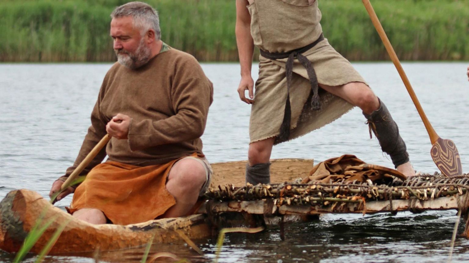 Two men, one seated, row a log boat across the River Nene