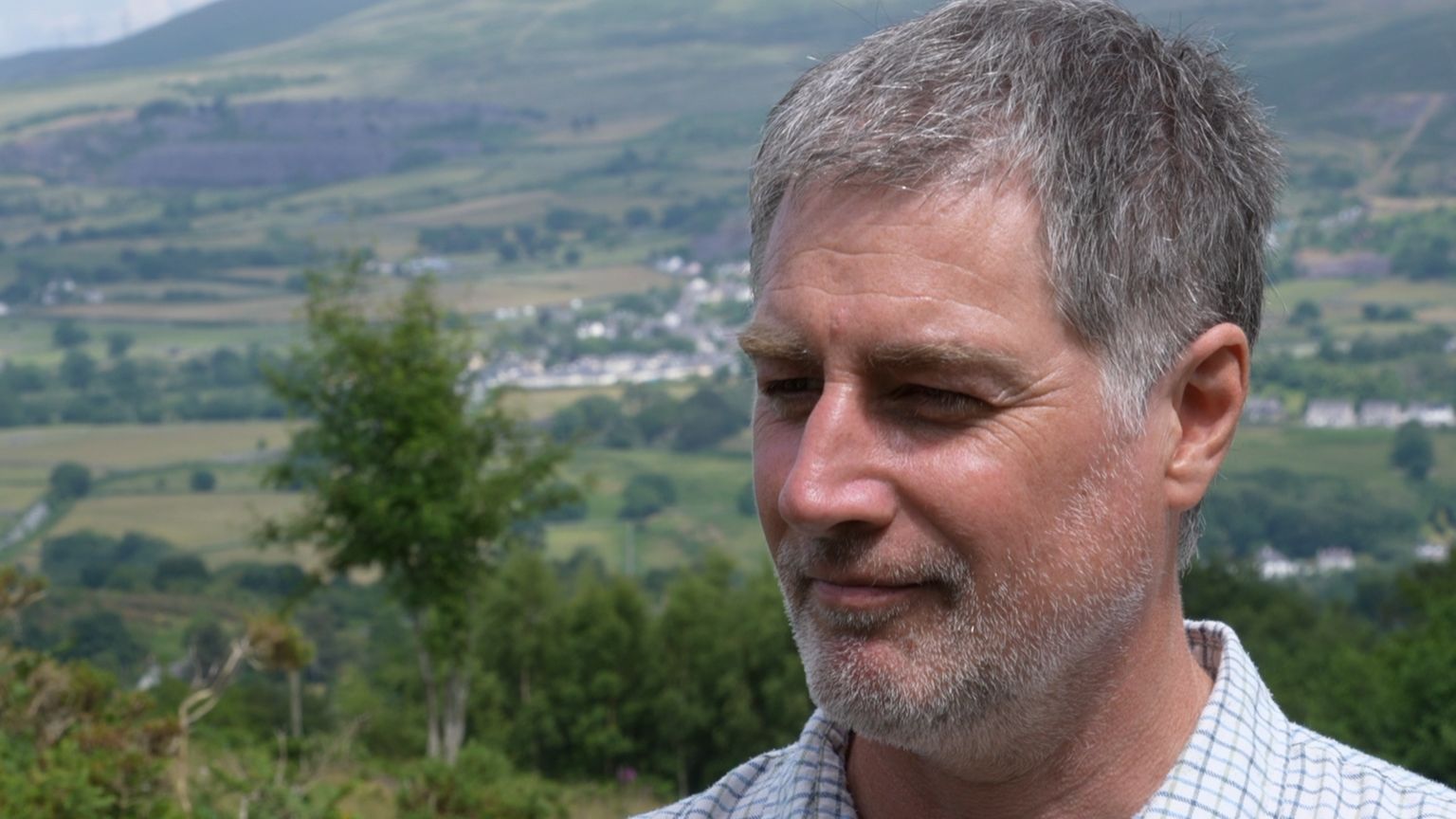 Head shot of Dr Craig Shuttleworth, outside, wearing pale checked shirt, with hillside and greenery in background