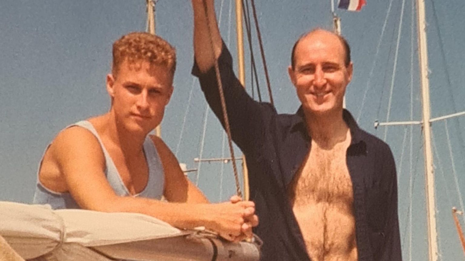 A young man standing with his older father on a yacht, leaning on the boom and smiling