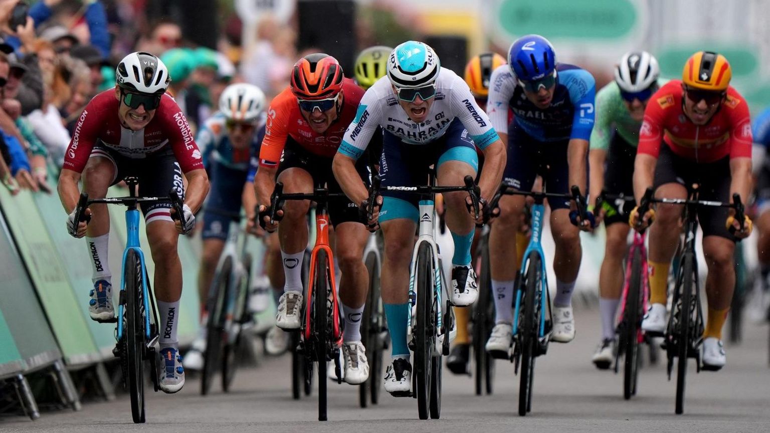 Nine cyclists wearing sunglasses and multi coloured outfits and helmets racing with gritted teeth.