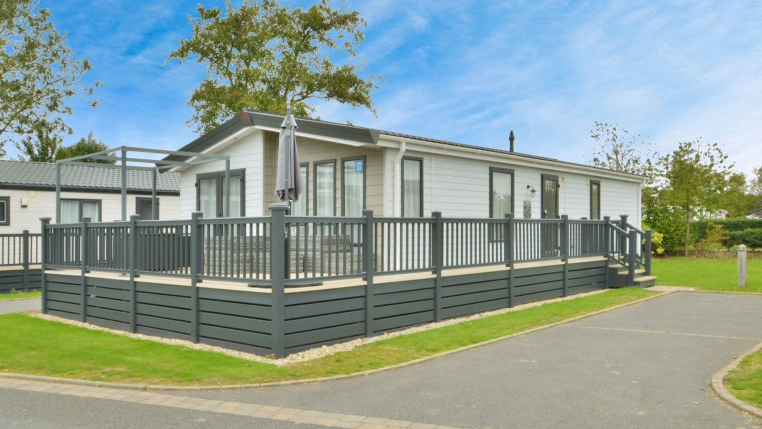 One of the lodges located at Cotswold Water Park. It is a one-storey building of a modern design with white wood panelling and a wooden fence and base, green in colour