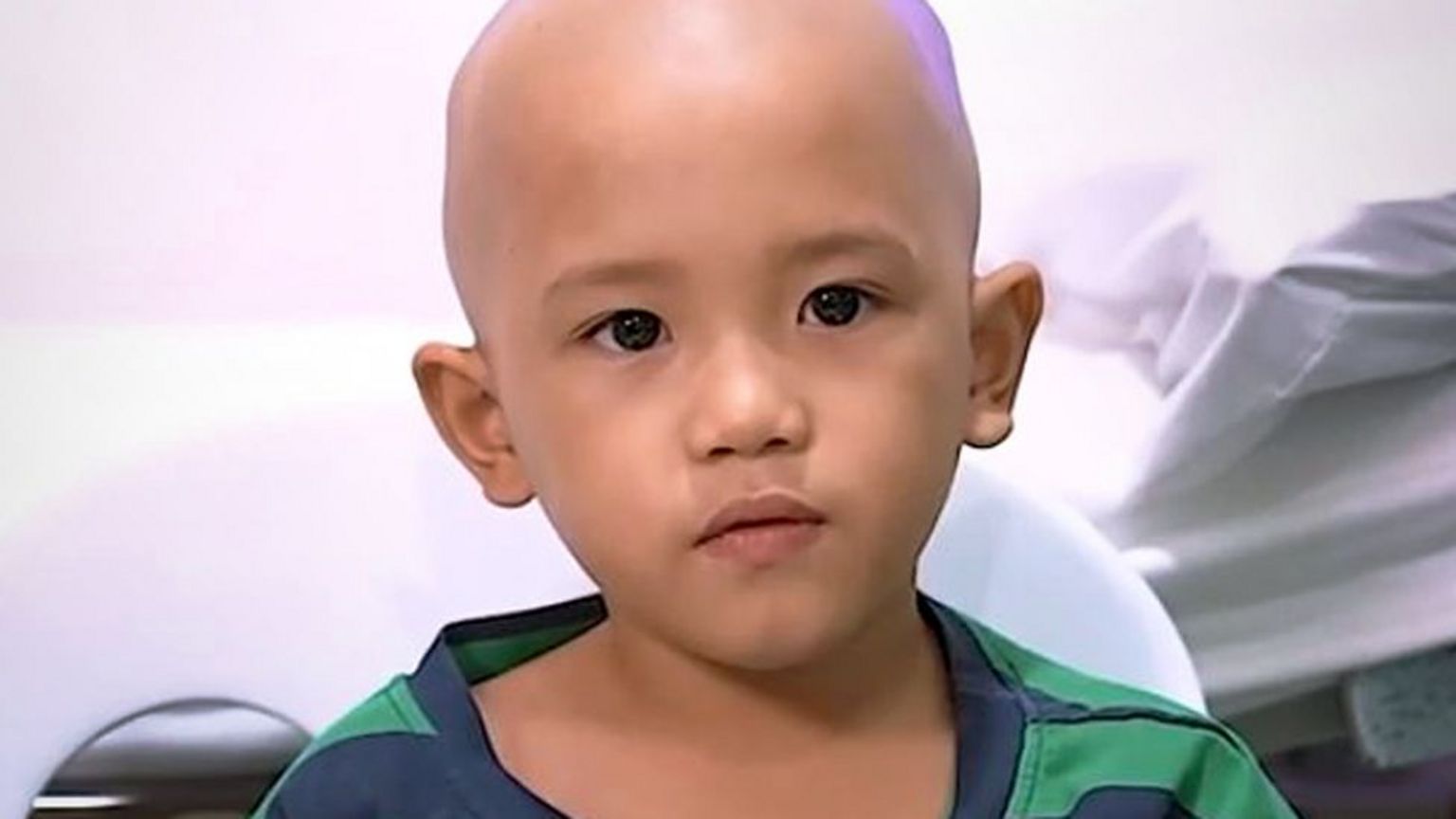 一名菲律宾男童卡利尔(Khalil)身穿绿蓝条纹T恤,剃着光头,上衣别着一个小型麦克风,背景为白色医院环境;他面向镜头,正在说话中