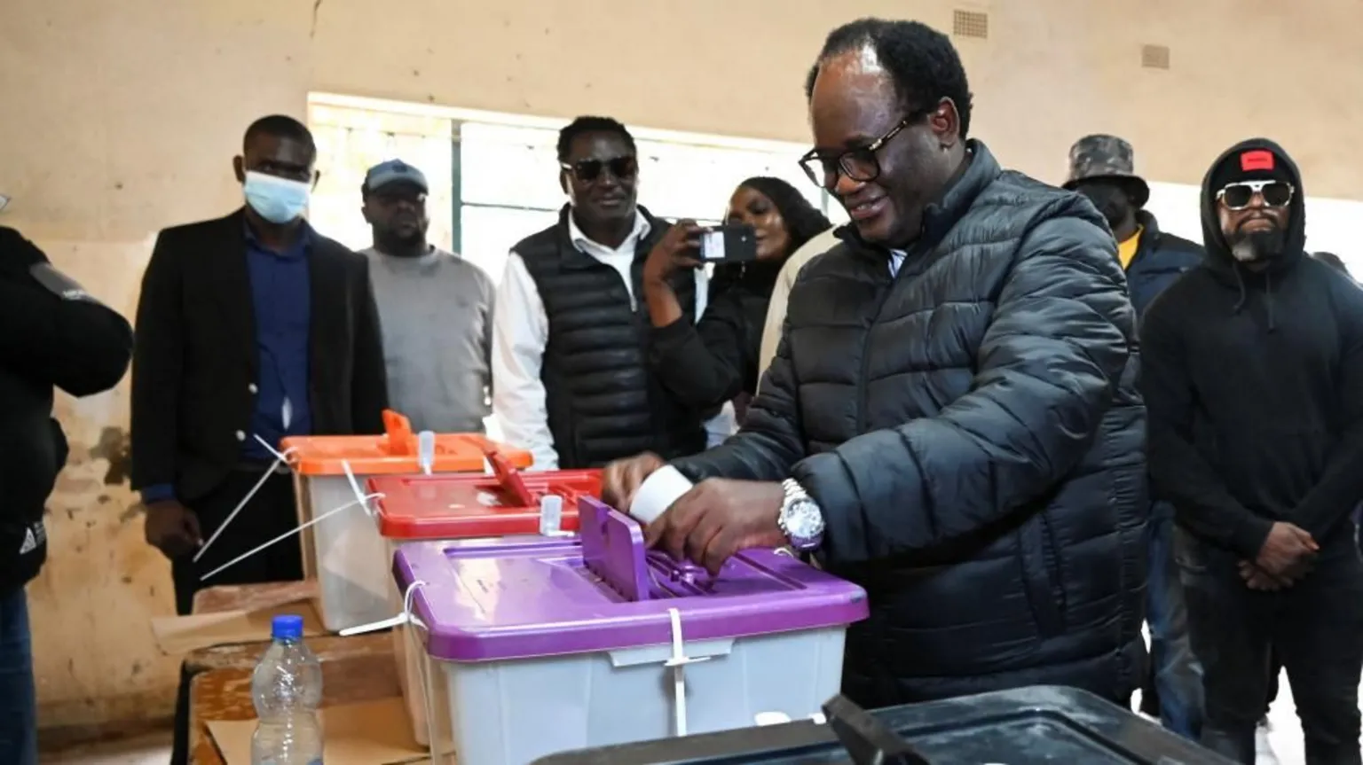 Reuters Brian Mundubile casts his ballot wearing a dark puffer jacket. He is placing his vote in a a ballot box with a purple lid. People are standing behind him watching what's going on.