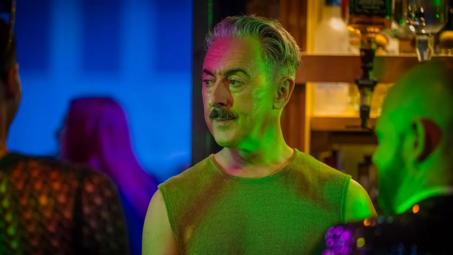 Channel 4 Actor Alan Cumming is in a night club, he's looking worried and is looking away to the left of the camera. The lighting in the bar is green and you can see bottles of alcohol in the background.