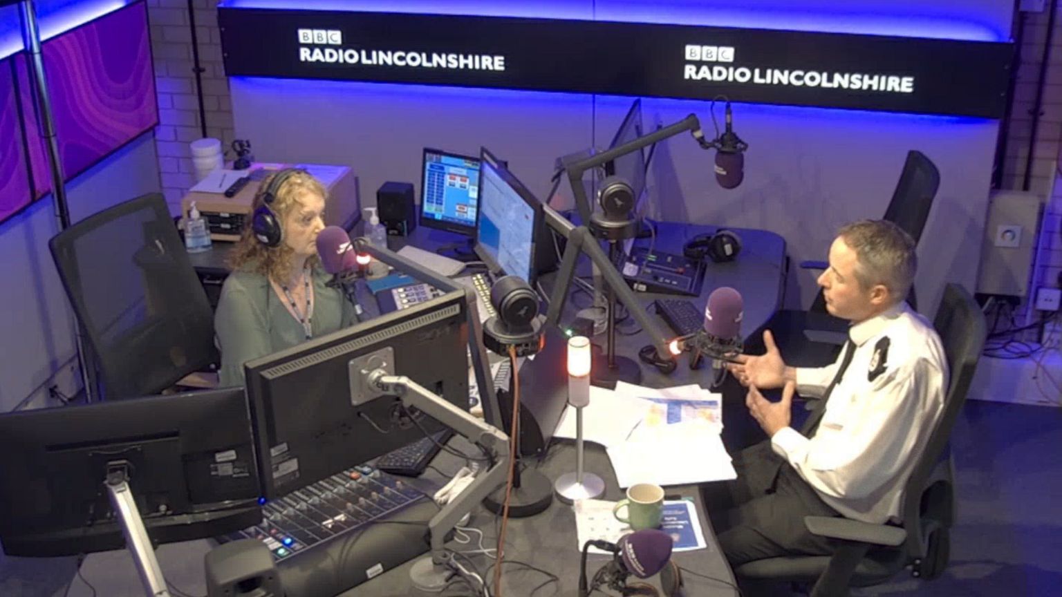 Chief Constable Paul Gibson in BBC Radio Lincolnshire's radio studio. He is photographed in his uniform in a purple studio, opposite presenter Frances Finn. Red lights on the microphones show it is live on air.