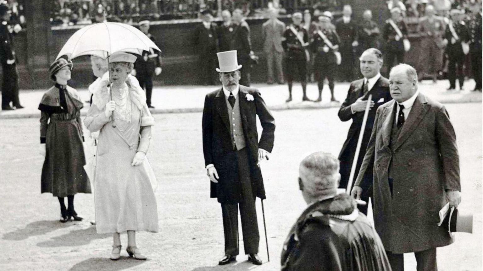 King George V and Queen Mary being welcomed by notable figures at the tunnel entrance. He is holding a cane and wearing a top hat and tails.  She has a white parasol and matching ornate dress. 