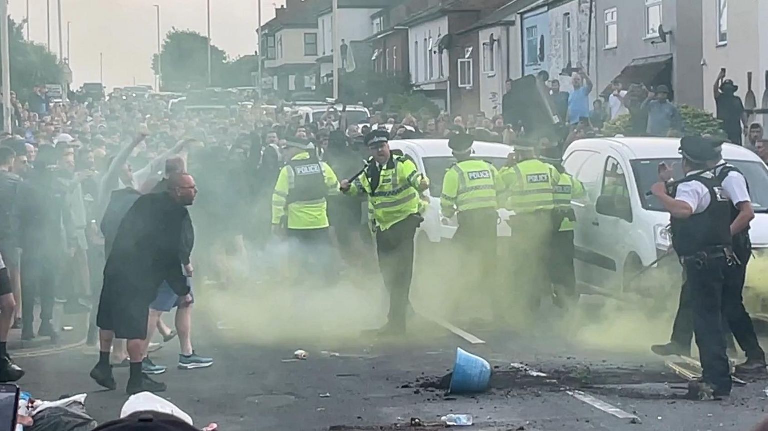 Hundreds of protesters gather in a street and are faced by a small number of police officers in hi-vis jackets and uniform holding batons