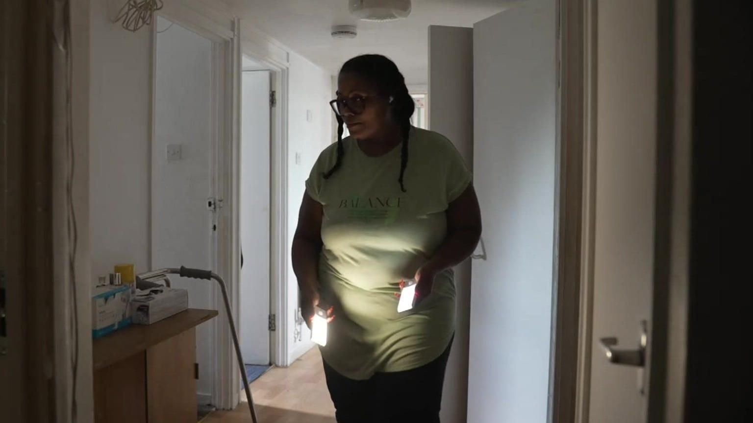 Ms Scott, a woman with dark hair in braids wearing a green t-shirt and black trousers, carring two portable LED lights through the dark hallway of her home