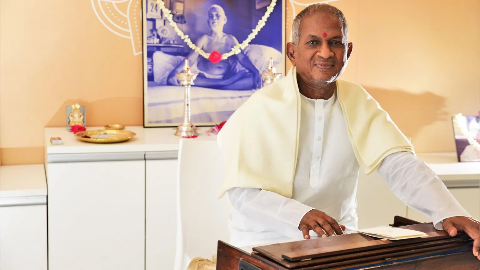 Ananda Vikatan Ilaiyaraaja sitting with a harmonium. He is wearing a white Indian kurta and an off white scarf.