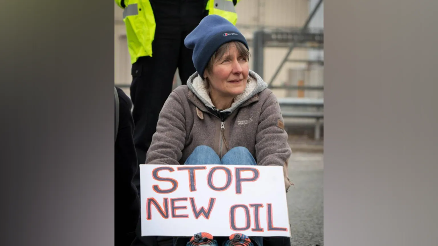 A woman sits on the ground wearing a blue beanie, gray hoodie, and jeans, holding a white protest sign that reads 