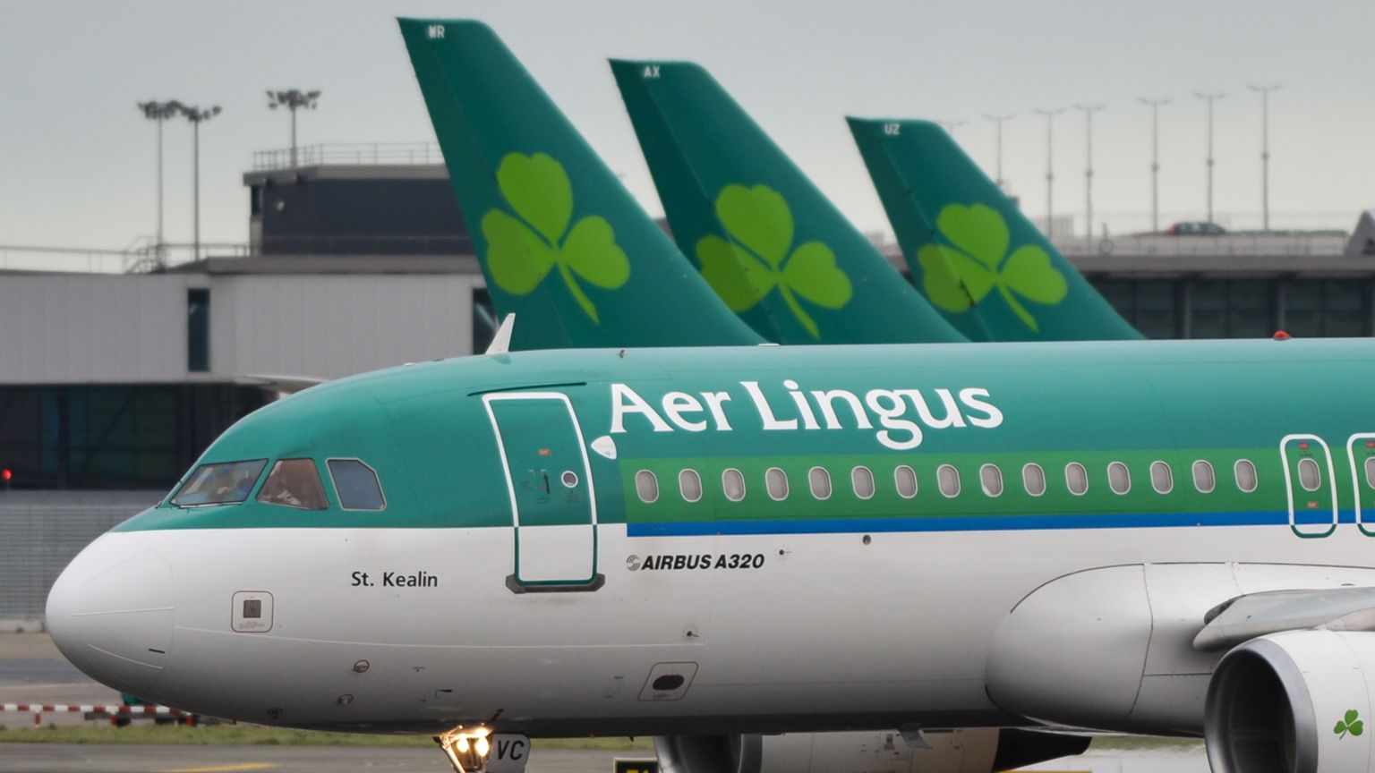 Green and white Plane with Aer Lingus written across it with three plane tail wings in the background that each have the Aer Lingus shamrock logo on them