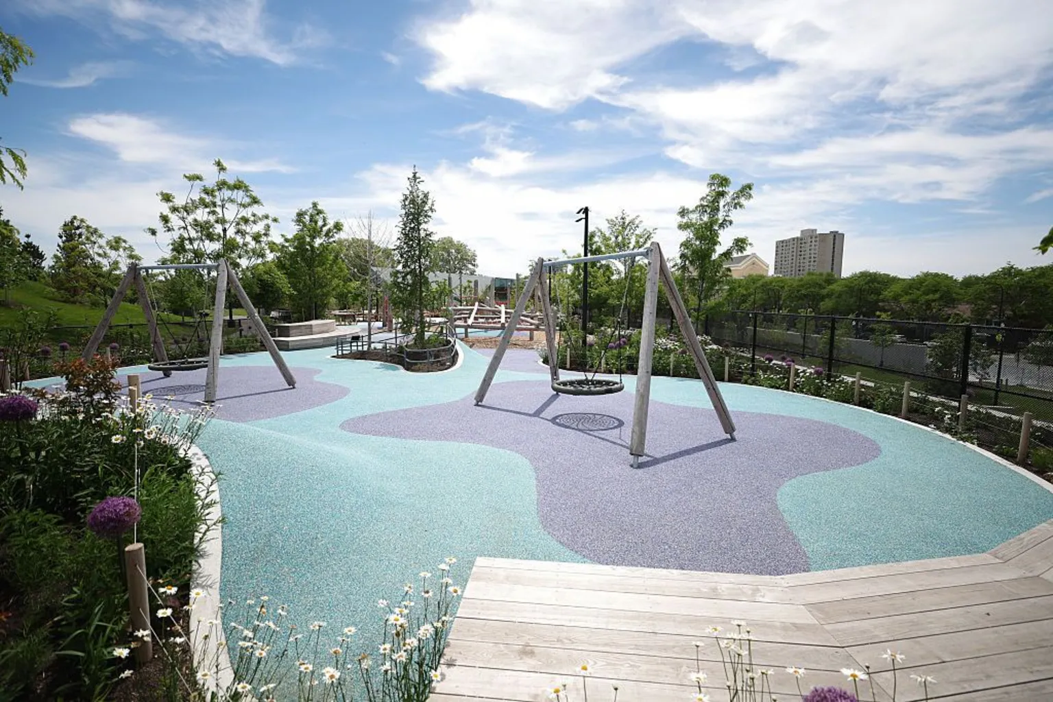 Talia Sprague/Bloomberg via Getty Images Two sets of swings are shown on a teal and purple playground surface. They are surrounded by lush landscaping and wooden benches and walking paths.
