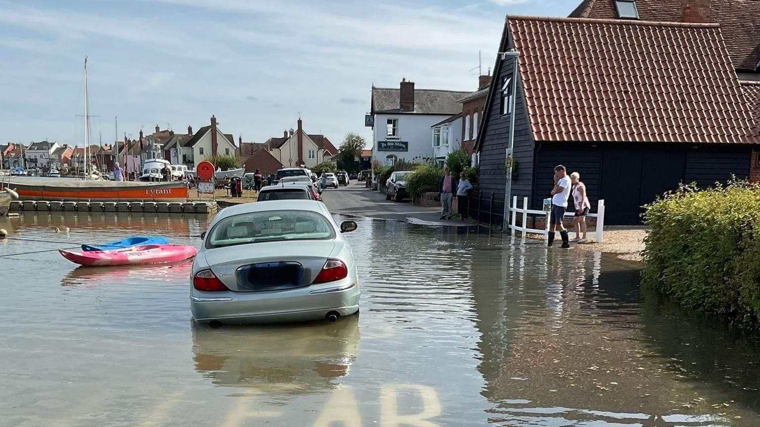 A car parked in the middle of a flooded road, with buildings to the right and the river to the left. 