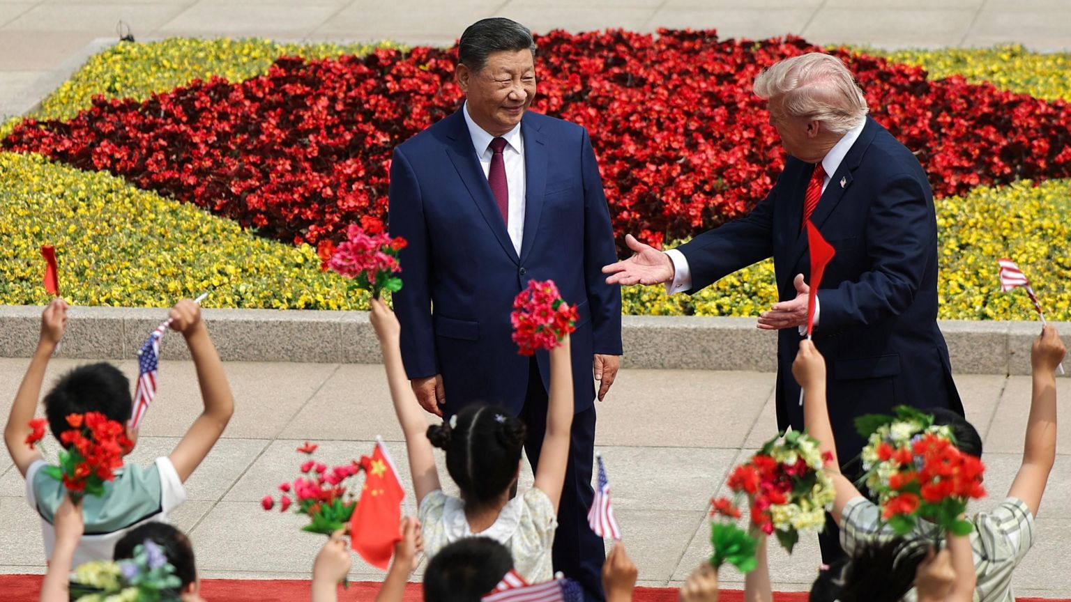 儿童在习近平和特朗普面前挥舞旗帜和鲜花，并举起手臂。习近平和特朗普站在红地毯上，特朗普向孩子们打手势。
