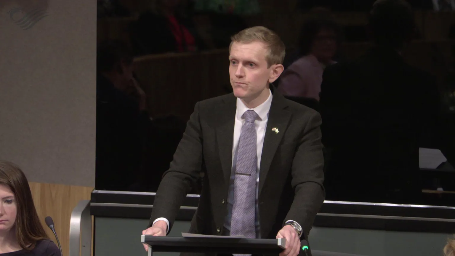 Senedd Cymru Joe Martin speaking at a lecturn in the Senedd, while a colleague sat to the left of him looks down.