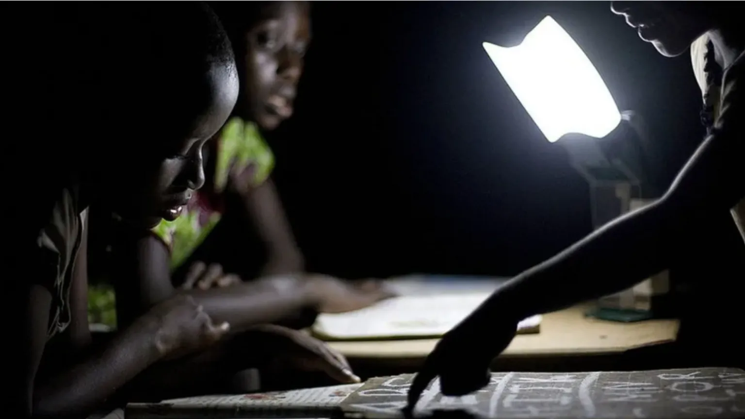 Getty Images Three children study by the light of a bright lamp in a dark room, with one child pointing at a page while the others lean over their books
