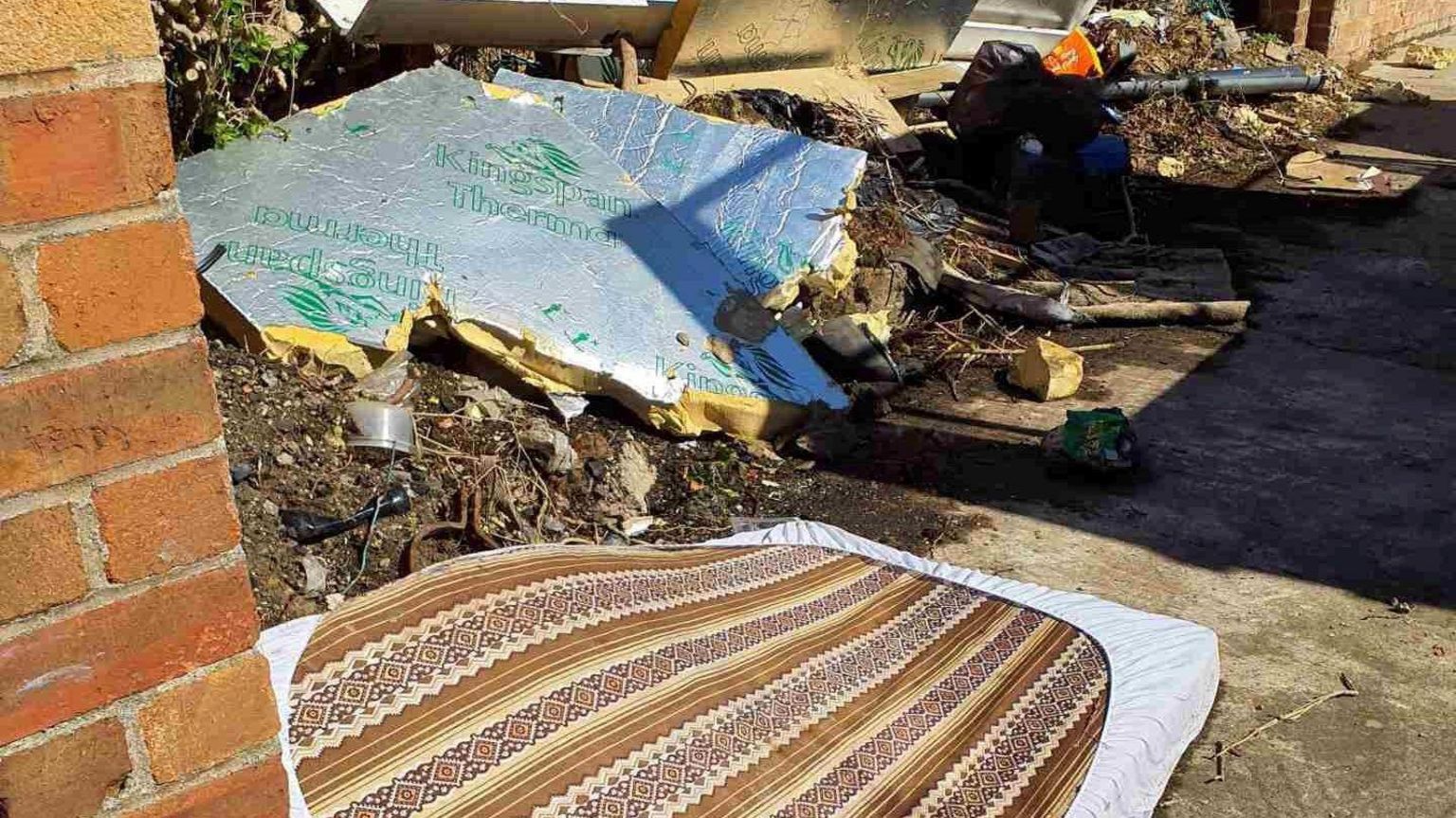 A brown and cream mattress has been dumped behind a wall next to broken insulated cladding panels.