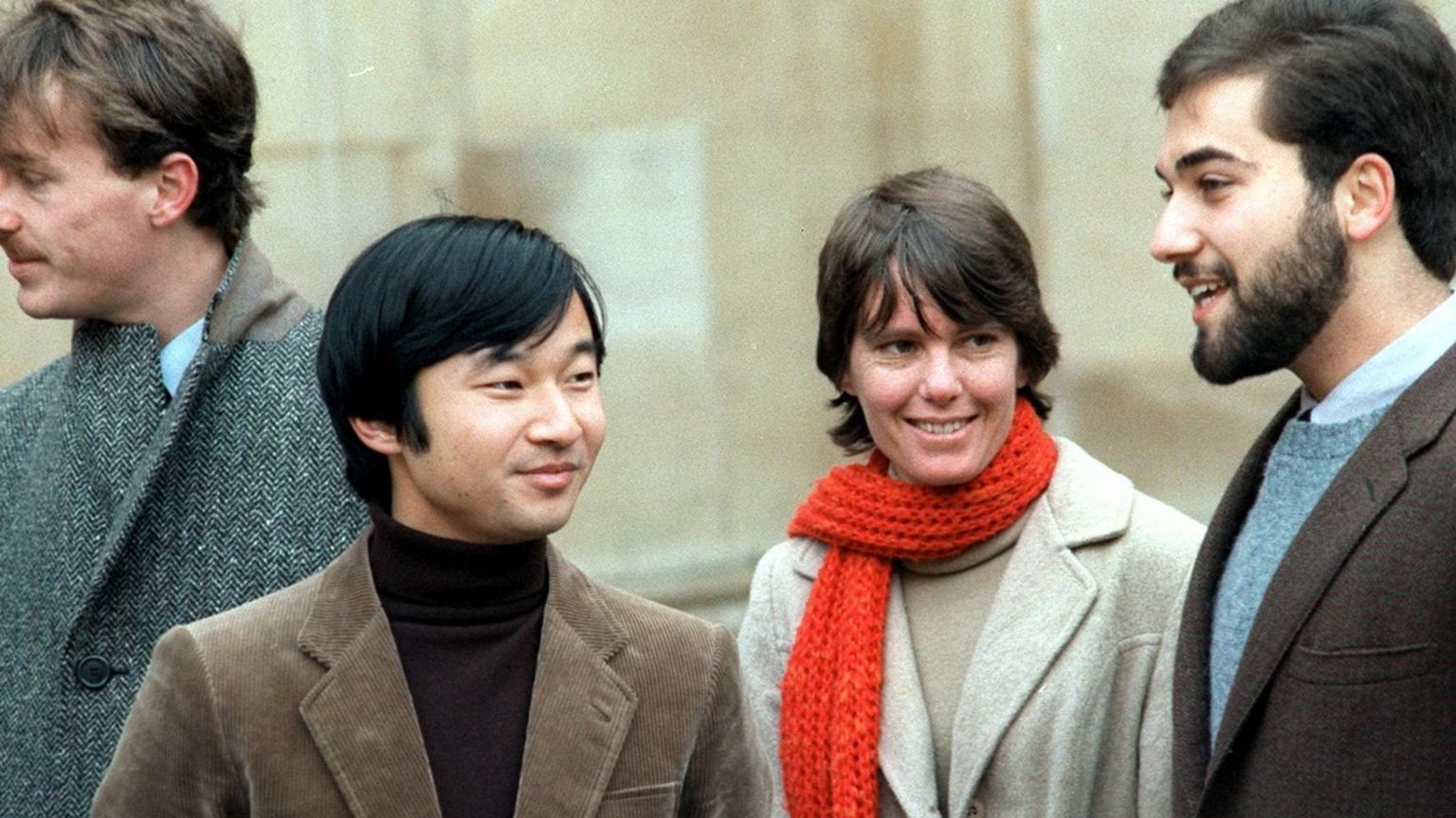 The Japanese emperor Naruhito as a student at Oxford, speaking to Keith George. The future emperor is smiling and wearing a black top, underneath a brown jacket. Keith George wears a blue top and a brown jacket. In the background, a man in a grey coat is looking away form the camera. A smiling woman with a red scarf around her neck looks at Keith George 