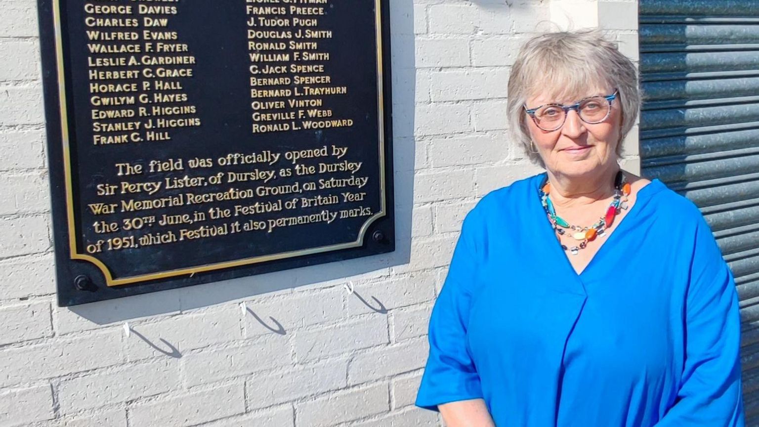 Julie Campbell wearing a blue shirt and stood next to a plaque with names of fallen soldiers on it