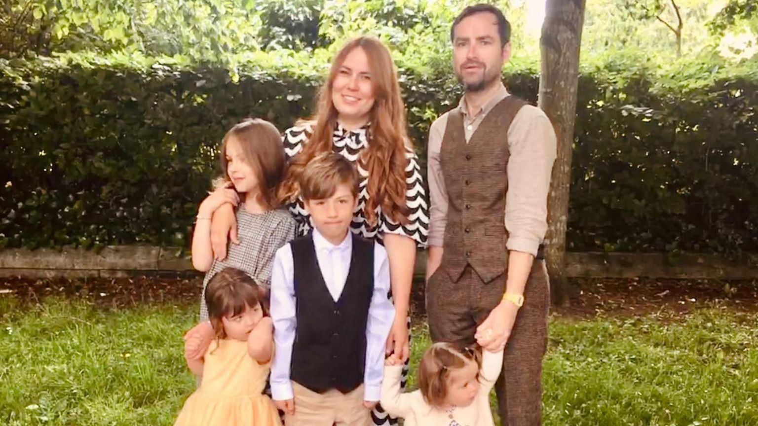 A family of six smiling at the camera. Liisa has her arm around one of her daughters, and Gwydion is holding one daughter's hand, are standing at the back while their four children are standing in front of them