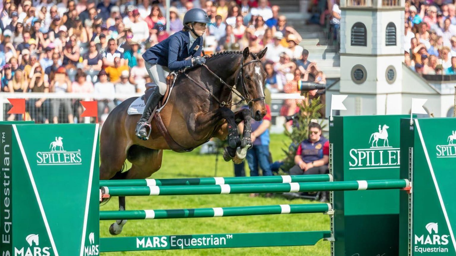 Alice Casburn on a horse clearing a fence in a show jumping course as a packed crowd watches on