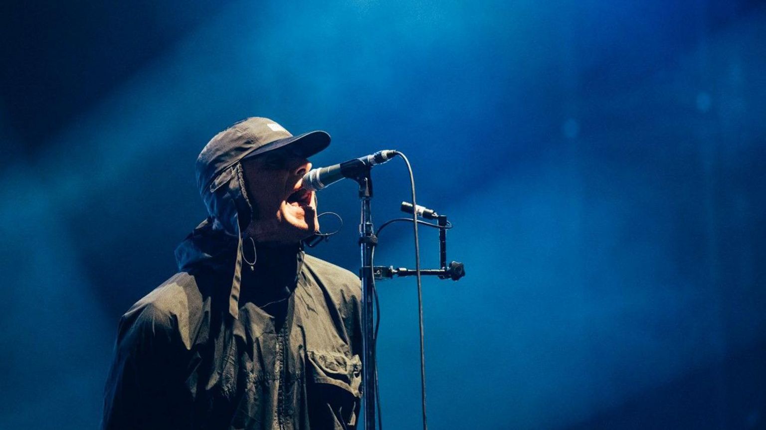 Liam Gallagher singing into a microphone at Reading Festival 2024. He is wearing a big coat and a peaked cap with flaps over the ears