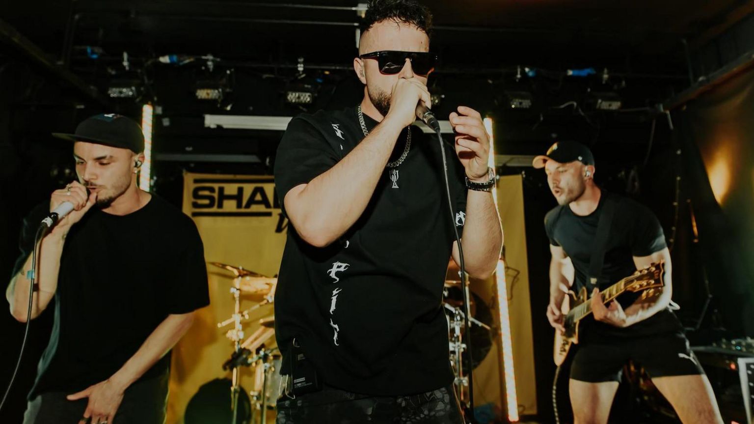 James Chakraverty (centre) performing on stage with his band Shade UK. There is a man singing into a microphone to the left and a man playing a guitar to the right. James Chakraverty is wearing a black t-shirt, dark black sunglasses and is wearing a silver necklace.