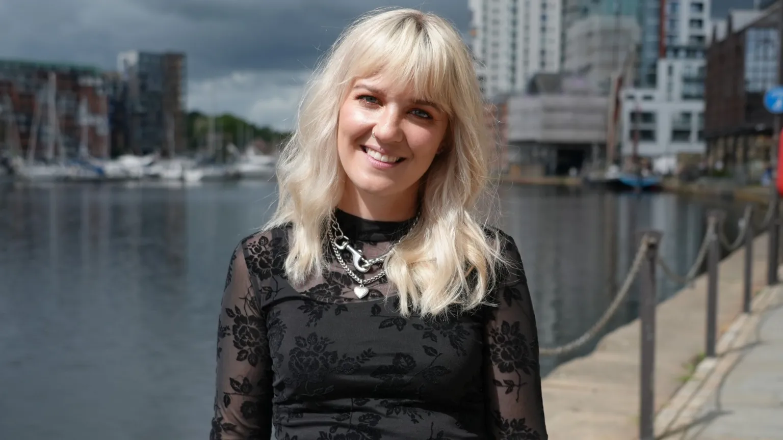Shaun Whitmore/BBC A woman with long blonde hair stands in front of a marina. She wears a black top with some floral patterns on it. She has silver chains around her neck.