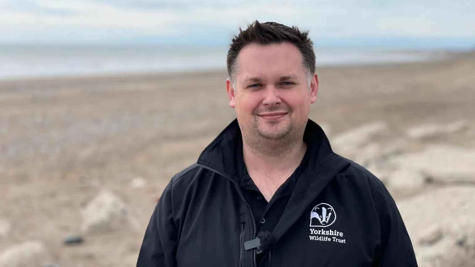 A man with short black hair, who is wearing a blue Yorkshire Wildlife Trust fleece, is standing on a sandy beach with the sea in the distance behind him
