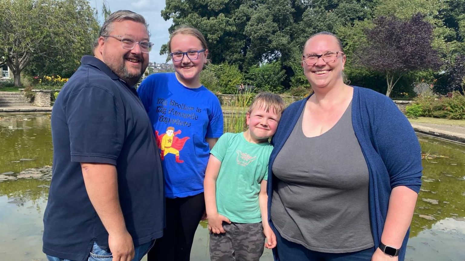 From left to right, Oli, Sophie, Toby and Nikki are stood in front of a park pond. There are trees in the background.  