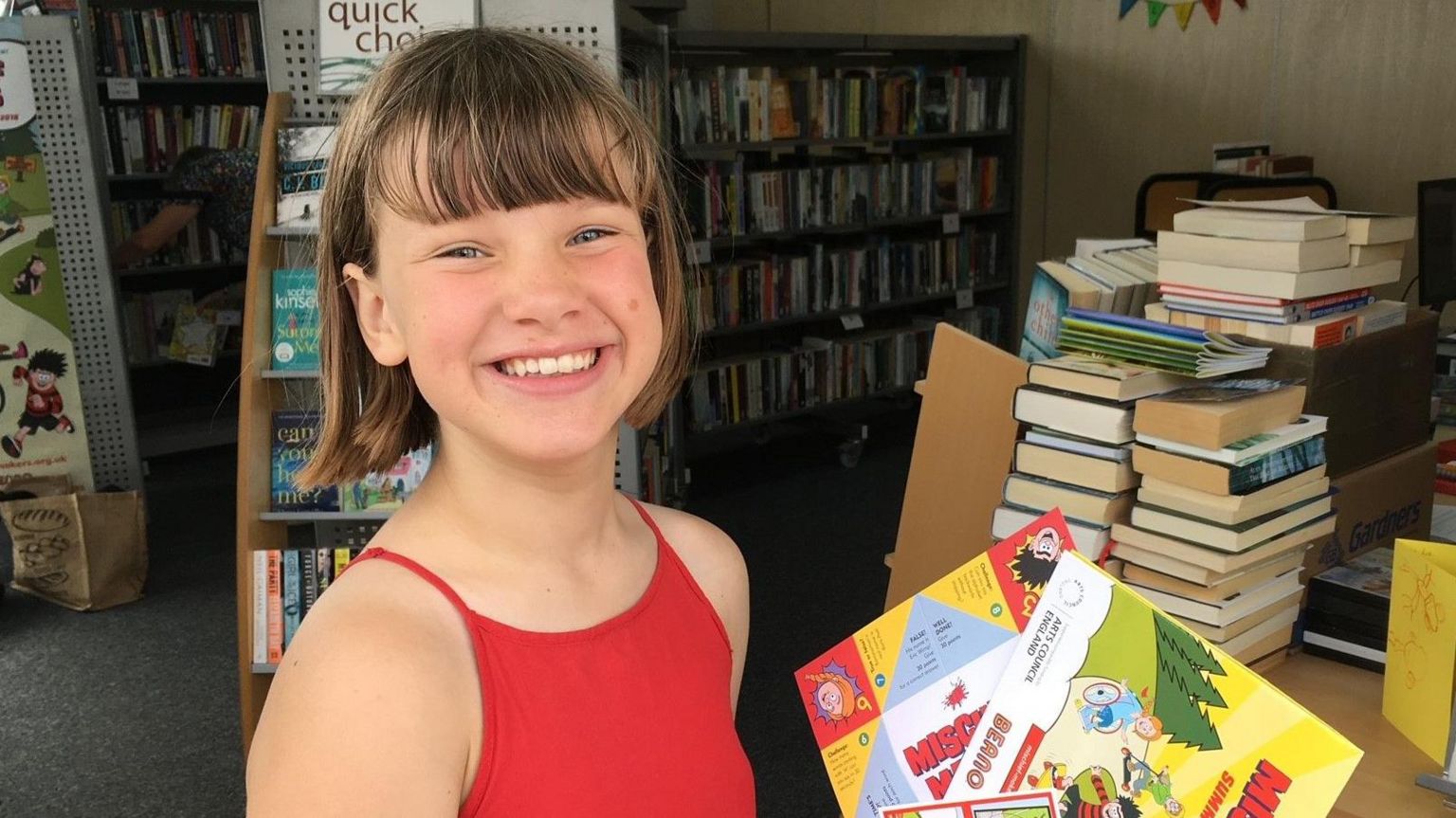 A young Wini is smiling in a red top, holding copies of comics at Ringmer Village Library in East Sussex. 