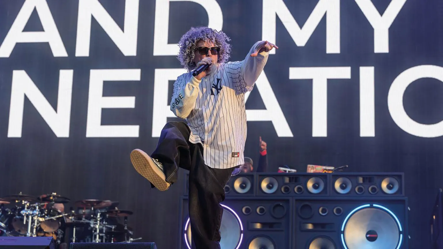 Getty Images A man wearing a white shirt and black trousers, singing into a microphone. He is pointing to the right with his left hand and kicking his right leg into the air.