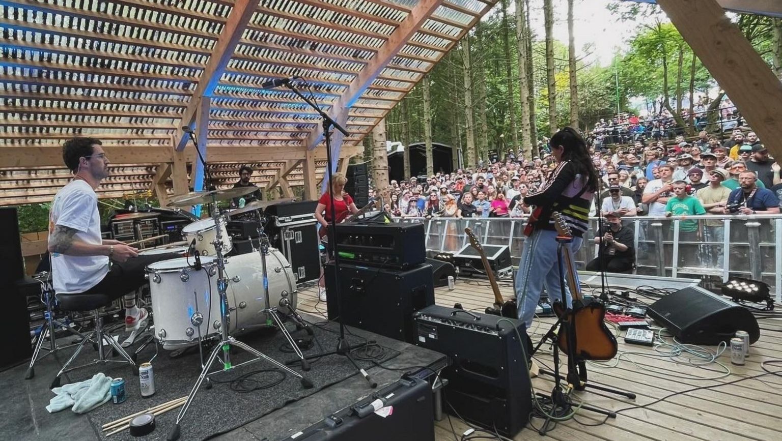 A woman plays a guitar on a stage, with a man playing drums and another woman playing guitar, in front of an outdoor crowd