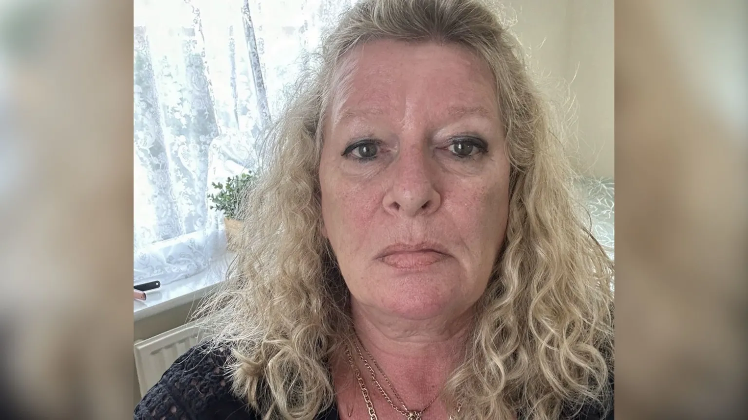 Laraine Chung A head and shoulders selfie-style photo of Laraine Chung. She is pictured in a lounge at home, with a house plant in a window behind her. She has blonde curly hair falling just below her shoulders and is wearing a black crocheted-style top and two gold necklaces. She is looking straight into the camera and is not smiling.