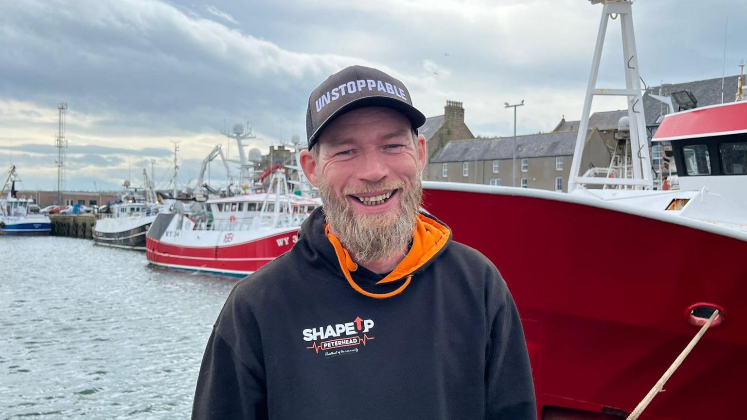 Man in a black hoodie and cap, smiling, standing in front of fishing boats at a dockside