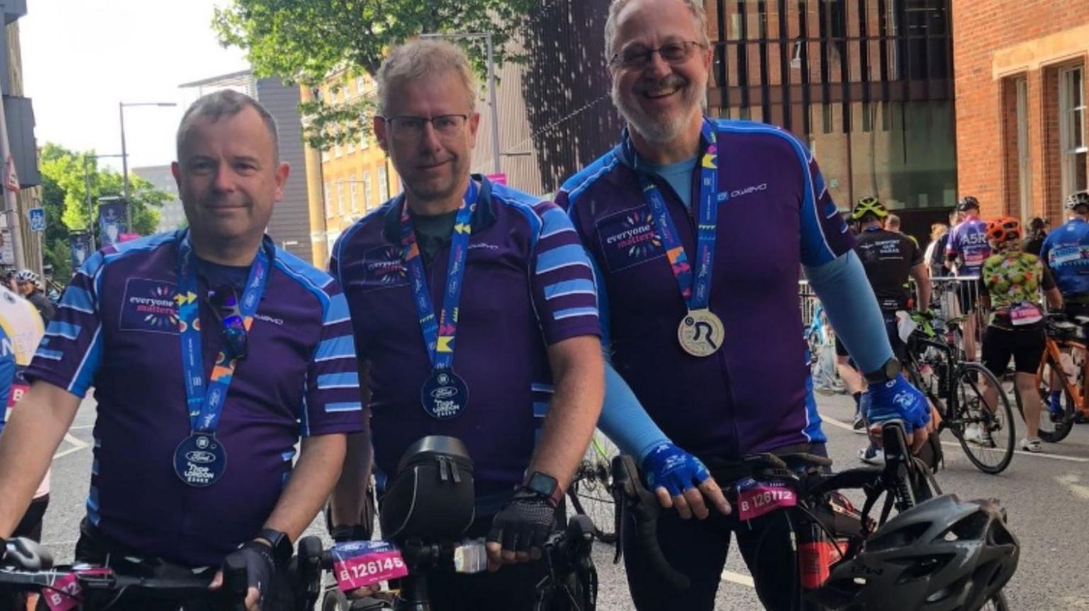 Three middle aged men in bright purple cycling jerseys stand beside their bikes wearing medals and smiling at the camera