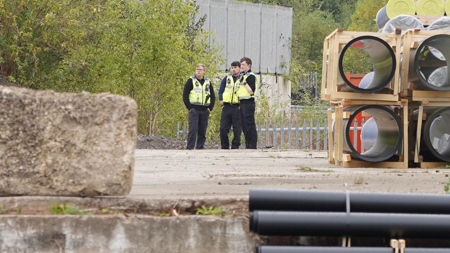 Three police officers standing on land at an industrial estate. They are all wearing police uniforms - high vis jackets and black trousers