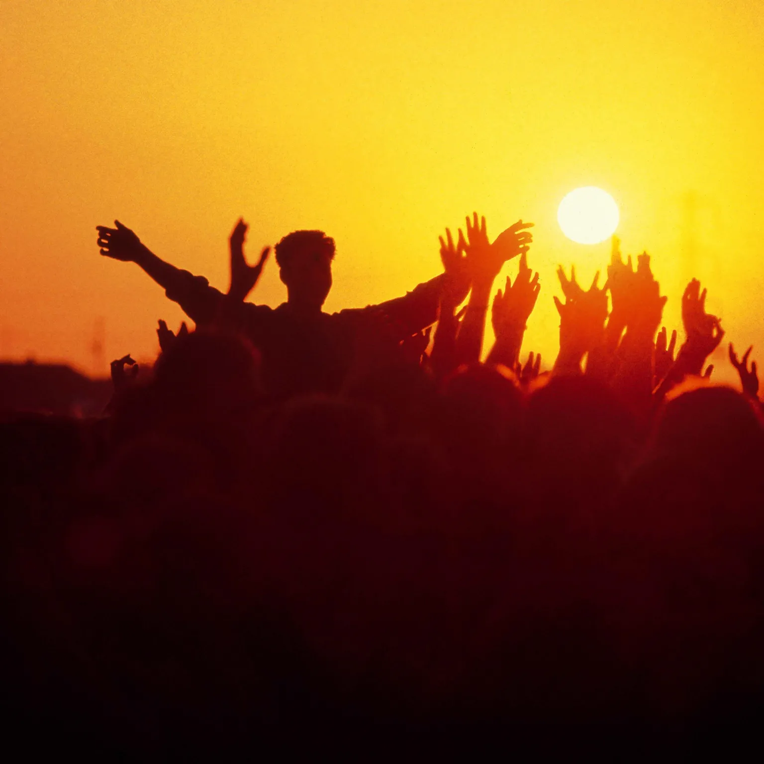 Mick Hutson/Redferns via Getty Images Crowds in silhouette holding arms in air in front of sunset, at Glastonbury Festival
